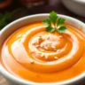 A small bowl of creamy Bang Bang Sauce, garnished with sesame seeds and cilantro, sitting on a rustic kitchen counter.