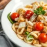 A close-up of Caesar pasta salad in a white bowl, showcasing pasta, lettuce, tomatoes, croutons, and Parmesan cheese with creamy dressing.