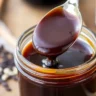 A close-up of a jar filled with thick, glossy gluten-free teriyaki sauce, with a spoon lifting the sauce to reveal its smooth consistency.