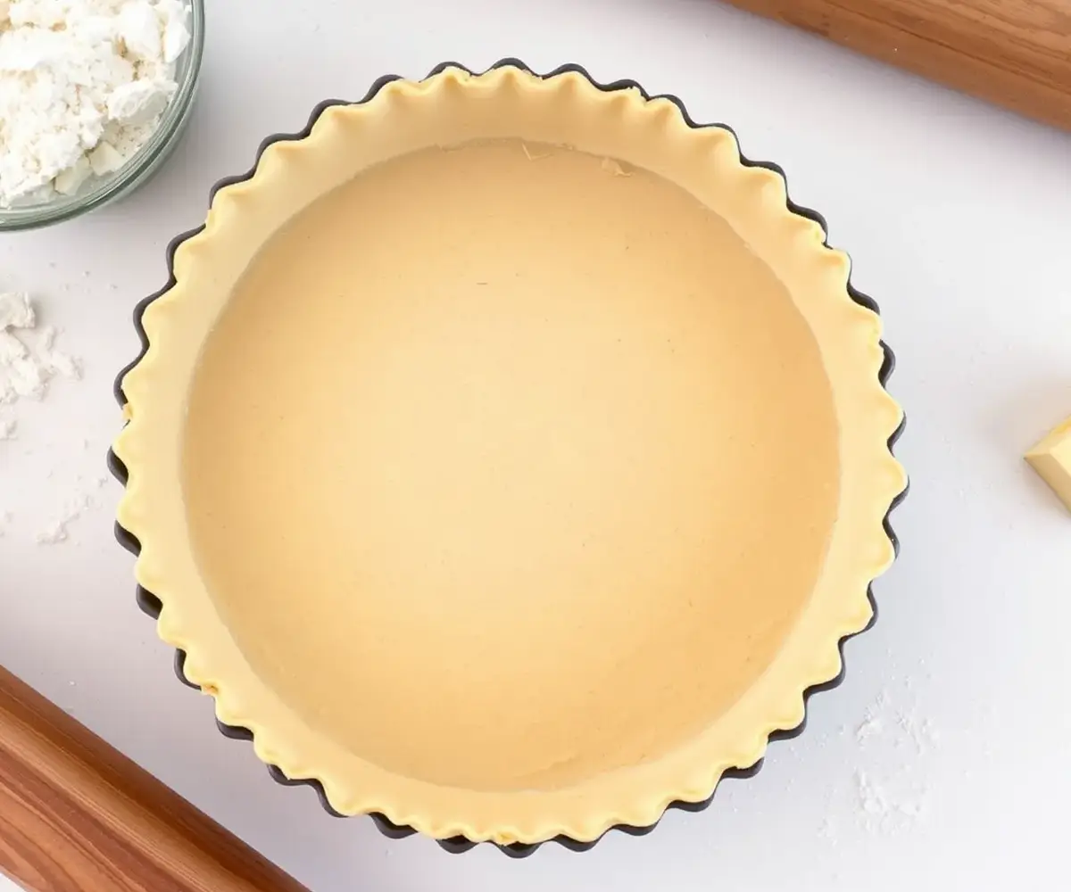 Golden-brown, freshly baked shortbread crust in a tart pan with visible flaky edges, surrounded by ingredients like butter, flour, and sugar.