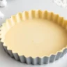 Golden-brown, freshly baked shortbread crust in a tart pan with visible flaky edges, surrounded by ingredients like butter, flour, and sugar.