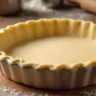 Golden shortbread crust recipe in a pie dish on a wooden kitchen countertop with kitchen tools in the background.