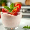 A creamy strawberry pudding served in a glass cup, topped with fresh strawberries and mint, with a soft-focus kitchen background.