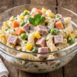 Receta de ensalada fría con pasta, jamón, zanahoria, elote y chícharos en un tazón de vidrio sobre una mesa rústica