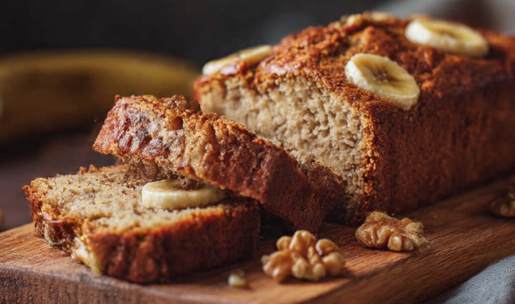 Ein detailreiches Food-Foto eines saftigen Bananenbrots ohne Zucker, aufgenommen in natürlichem Licht. Das goldbraune Bananenbrot liegt auf einem Holzbrett, umgeben von reifen Bananen und einer Tasse Kaffee. Ideal als Titelbild für den Blogbeitrag „Saftiges Bananenbrot ohne Zucker – Gesundes Rezept“ auf www.cucinarecipes.com .