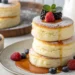 Stack of three tall fluffy Japanese soufflé pancakes with melting butter and maple syrup drizzling down the sides on a white plate