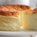 Fluffy Japanese cheesecake with golden top on white cake stand showing jiggly cotton-soft texture with slice being lifted