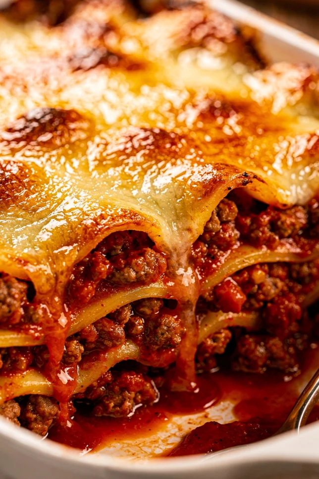 Freshly baked million dollar lasagna in a white ceramic dish with golden bubbly mozzarella on top and rich red meat sauce on the sides